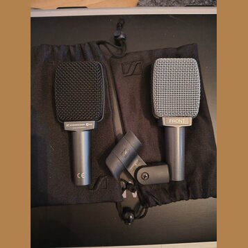 Dvě mikrofony s mřížkou a jeden držák v černých látkových pytlích. Levý mikrofon je černý s nápisem SENNHEISER e606 a MADE IN GERMANY, v dolní části…