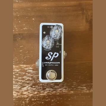 Kompresorový pedál s bílým rámem a černým horním panelem. Na panelu je bílý nápis „SP compressor BY xotic usa“. Pedál má dva průhledné ovládací knofl…