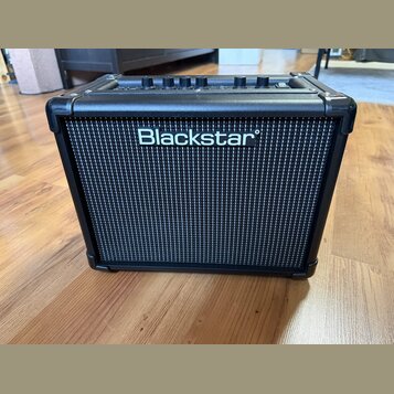 Černý kytarový kombo zesilovač s černou textilní mřížkou. Uprostřed mřížky je bílý nápis Blackstar. Na horní straně jsou vidět ovládací knoflíky a úc…