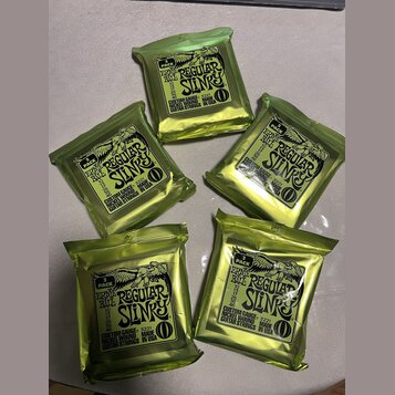 Pět uzavřených balení kytarových strun v zelených fóliových sáčcích. Na každém balení je černý štítek s nápisy ERNIE BALL REGULAR SLINKY, CUSTOM GAUG…