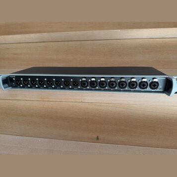 Šedý rackový audio přístroj s černým horním krytem. Na zadním panelu jsou umístěny konektory XLR a dva konektory Ethernet s nápisy SEND a RETURN. V l…