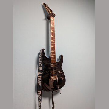 Černá elektrická kytara s hnědým hmatníkem a dvěma humbuckery. Na hlavě nástroje je viditelné logo Schecter. Kytara má nasazený černobílý popruh s ná…