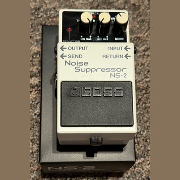 Bílý efekt pedál s černým spínacím tlačítkem a nápisem BOSS. Na přední straně je text Noise Suppressor NS-2 a označení vstupů OUTPUT, INPUT, SEND a R…