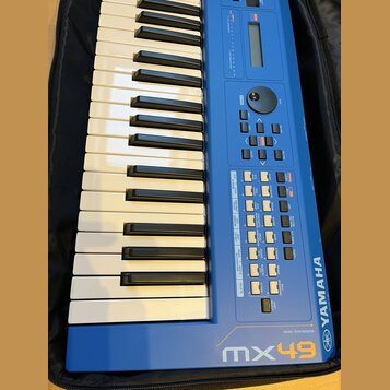 Modrý syntezátor s klaviaturaí, umístěný v černém pouzdře. Na pravé straně je svisle napsáno YAMAHA a v dolní části nápis MUSIC SYNTHESIZER mx49 spol…