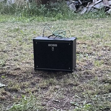 Černý kytarový kombo zesilovač s hranatým tvarem, umístěný na trave. Na přední straně je bílý nápis BOSS a pod ním symbol připomínající stylizované p…