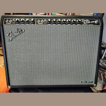 Kytarový zesilovač s černým krytím a šedou textilní mřížkou. Na horním ovládacím panelu jsou knofíky a nápisy NORMAL, VIBRATO, Twin Reverb-Amp. Na př…