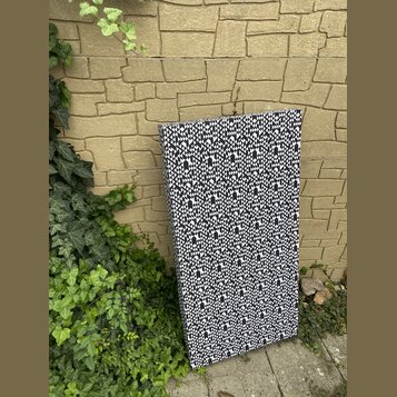 Obdélníkový akustický panel potažený látkou s černobílým geometrickým vzorem. Panel je opřen o žlutou kamennou zeď vedle zelené vegetace. Na povrchu…