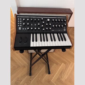 Černý syntetizátor s dřevěnými bočními panely umístěný na černém skládacím stojanu. Na ovládacím panelu je bílý nápis SUBSEQUENT 25 a logo Moog. Nást…