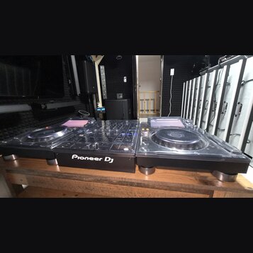 Sada DJského vybavení v černé barvě složená ze dvou kontrolérů s jogovými koly a centrálního mixeru. Na přední straně mixeru je bílý nápis Pioneer DJ…