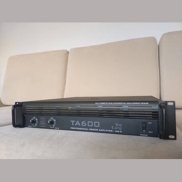 Černý rackový výkonový zesilovač s úchyty po stranách. Na předním panelu je bílý nápis TA600, logo the t.amp a text PROFESSIONAL POWER AMPLIFIER - MK…