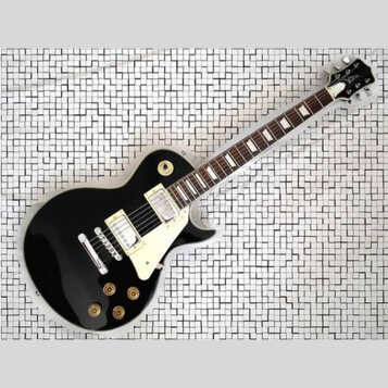 Černá elektrická kytara s bílým pickguardem a dvěma humbuckery. Na hlavě je viditelné logo Gibson. Nástroj vykazuje známky běžného používání, povrch…