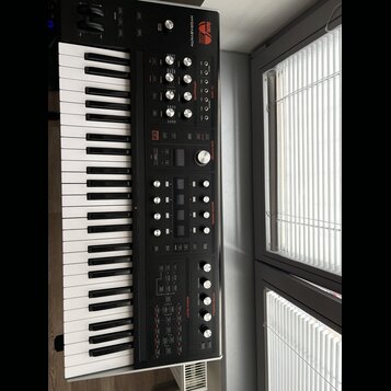 Černý synthesizér s klaviaturaí, na jehož horním panelu je bílý nápis HYDRASYNTH a logo MOOG. Přístroj obsahuje množství ovládacích knoflíků, posuvní…