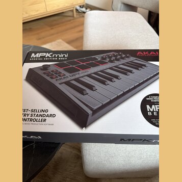 Fotografie zachicuje krabici s produktem AKAI MPKmini SPECIAL EDITION GREY. Na obalu je zobrazen šedý kompaktní MIDI kontrolér s klávesami, osmi červ…