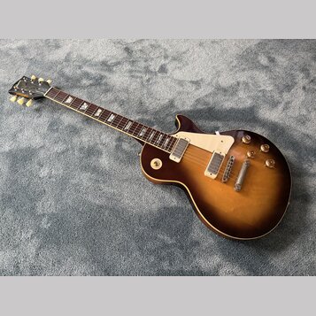Elektrická kytara s tónem sunburst v hnědých odstínech a bílým pickguardem. Na hlavě je viditelné logo Gibson. Hриф má tmavý hmatník s trapezoidovými…