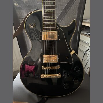 Černá elektrická kytara s gold hardwarem a krémovým lemováním. Na těle jsou dva humbuckery, čtyři ovládací knofíky a přepínač s nápisy RHYTHM a TREBL…