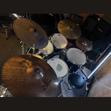 Celá bicí souprava s černými kotly a kovovými obručmi. Na činelech jsou viditelné nápisy Zildjian, ZBT a VFIP. Na bubnech je vidět logo TAMA. Souprav…