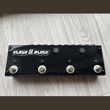 Černý obdélníkový pedalboard s nápisem ULASA & BLACK v bílém stylizovaném písmu. Na přední straně jsou čtyři kovové spínací tlačítka s bílými podložk…