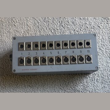Šedý kovový box s dvěma řadami deseti XLR konektorů, celkem 20 kusů. Horní řada je neoznačená a spodní řada je popisaná čísly 1 až 10. V levém dolním…