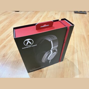 Černá cardboardová krabice s červenými prvky a úchytkou v horní části. Na přední straně je bílé logo a nápis AUSTRIAN AUDIO, fotografie sluchátek a v…
