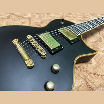 Černá elektrická kytara s matným povrchem a zlatým hardwarem. Na těle jsou dva humbuckery, kobylka a dva ovládací knoflíky v zlatém provedení. Na kyt…