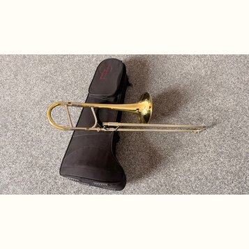 Zlatý mosazný trombon položený vedle černého měkkého pouzdra. Na pouzdře je v horní části červené kulaté logo s nápisem TENOR. Nástroj vykazuje v hor…