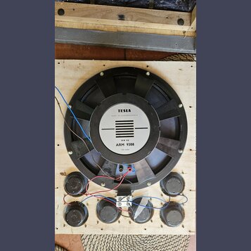 Na dřevěné desce je umístěn velký černý reproduktor s magnetem. Na středovém bílém štítku je nápis TESLA, MADE IN CZECHOSLOVAKIA, 50 W-8Ω a ARM 9308,…