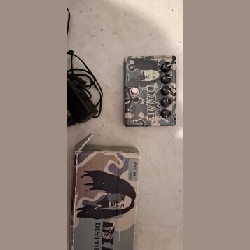 Kytarový efekt pedál s kamuflážním vzorem a černobílým portrétem osoby. Na pedálu je nápis DIME DISTORTION a logo MXR. Vedle pedálu je černý napájecí…