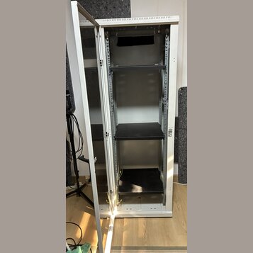 Bílá kovová serverová skříň s otevřenými dveřmi. V interiéru jsou umístěny tři černé police na různých úrovních. Povrch skříně vykazuje běžné známky…