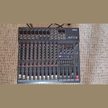 Šedý mixovací pult s nápisy YAMAHA a Mx12/4. Zařízení obsahuje několik vstupních kanálů s posuvníky, knoflíky a červenými kontrolkami. V pravé části…