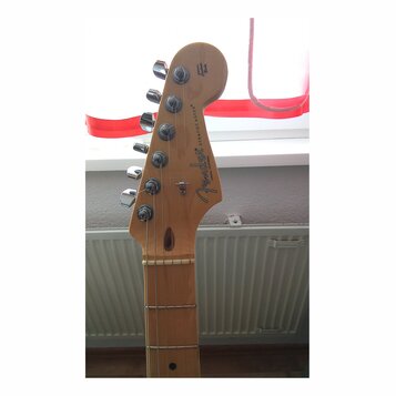 Fotografie zachycuje hlavu elektrické kytary v přírodním dřevě s chromovanými ladičkami. Na hlavě je černý nápis Fender, STRATOCASTER® a menší text C…