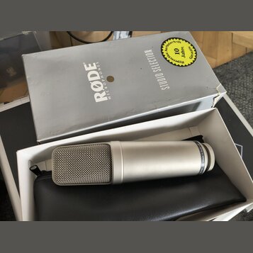 Stříbrný kondenzátorový mikrofon s kovovou síťkou, na jehož těle je nápis Made in Australia. Mikrofon leží v bílém boxu na černém koženěném pouzdře.…