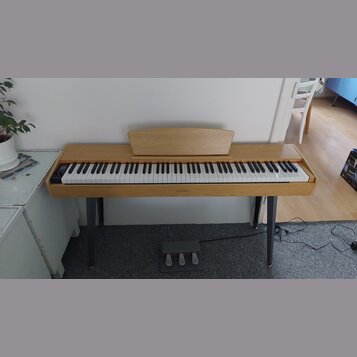 Digitální piano v dřevěném provedení s bílými a černými klávesami. Na středové liště pod klaviaturou je nápis HENRY'S. Nástroj stojí na čtyřech šedýc…