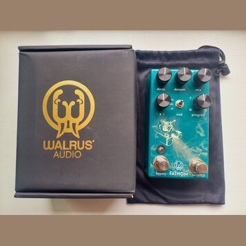Na fotografii je tyrkysový kytarový efekt v černém textilním vakle a vedle něj černá krabice s goldeným logem a nápisem WALRUS AUDIO. Na efektu jsou…