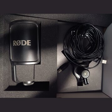 Černý mikrofon s nápisem RØDE umístěný v originální pěnové výplni. Vedle něj je srolovaný černý kabel USB a držák mikrofonu. Všechny komponenty vypad…