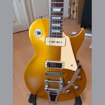 Zlatá elektrická kytara s jednodrážkovým tělem a bílým plechtrzem. Nástroj je vybaven dvěma humbuckery, chromovaným mostem s tremolo páčkou a čtyřmi…