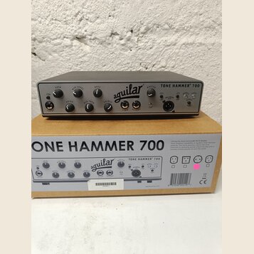 Šedý basový předzesilovač s nápisem TONE HAMMER 700 a logem aguilar. Zařízení má několik ovládacích knoflíků s popisky GAIN, MID LEVEL, BASS, DRIVE,…