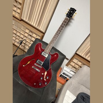 Polodupatá elektrická kytara v červené barvě s černým pickguardem. Na hlavě hmatníku je viditelné logo Gibson. Kytara má dva humbuckery, tři ovládací…