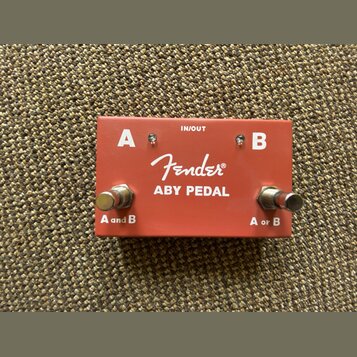 Červený kovový kytarový pedál s nápisem Fender ABY PEDAL. Na horní straně jsou bílá písmena A a B, nápis IN/OUT a dva kovové spínače s popisky A and…