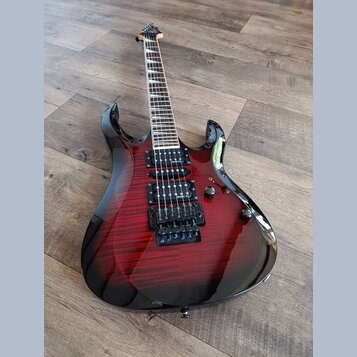 Elektrická kytara s červeným gradientním lakem a viditelnou strukturou dřeva. Nástroj má dva humbuckery, hmatník s vložkami a černý most. Na kytaru j…