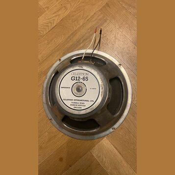 Kruhový reproduktor s kovovým rámem a magnetem. Na bílém štítku je uveden nápis CELESTION G12-65, IMPEDANCE 15 OHMS a adresa CELESTION INTERNATIONAL…