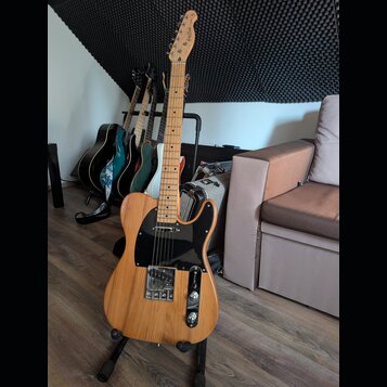 Elektrická kytara s přírodním dřevěným povrchem, černým pickguardem a chromovanými komponenty. Na hlavě je viditelné logo "Telecaster". V pozadí stoj…