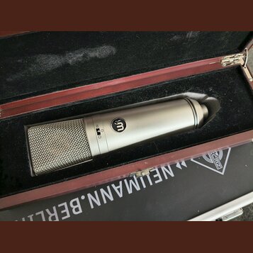 Kondenzátorový mikrofon stříbrné barvy v černé polstrované přepravní kufře. Na těle mikrofonu je viditelné logo „m“ v kroužku a přepínač s označením…