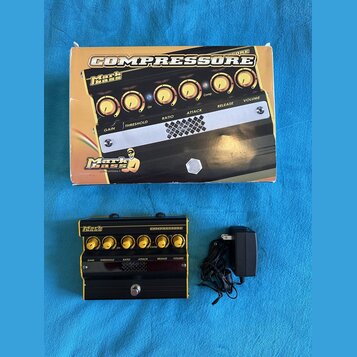 Fotografie zobrazuje kompresor Markbass COMPRESSORE v originálním kartonovém obalu a s přívodním adaptérem. Zařízení má černé tělo se zlatými ovládac…