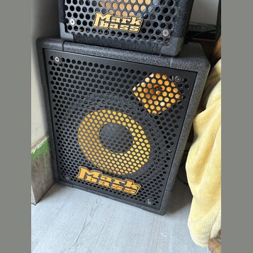 Černý basový reproduktor s kovovou mřížkou, na které je žlutý kruh a čtverec. V dolní části mřížky je umístěno žluté logo Mark bass. Nad ním je vidět…
