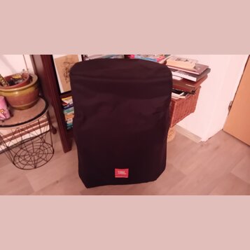 Černý textilní kryt na reproduktor nebo subwoofer. V dolní části je umístěno červené obdélníkové logo s bílým nápisem JBL. Povrch látky je v dobrém s…