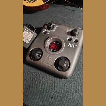 Šedý kytarový efekt pedál s nápisem GUITAR EFFECTS PEDAL G1 a logem ZOOM. Zařízení má dvě stoupací pedály, několik ovládacích knoflíků a červený digi…
