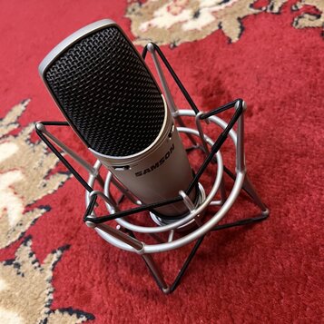 Stříbrný kondenzátorový mikrofon s černou mřížkou, umístěný v odpruženém držáku (shock mount) stříbrné barvy. Na těle mikrofonu je černý nápis SAMSON…