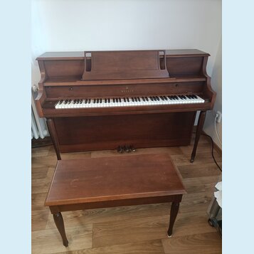 Hnědé dřevěné piano s doprovodnou stoličkou. Nad klávesnicí je vyveden nápis WILLIS. Povrch nástroje i stoličky vykazuje drobné známky opotřebení.
