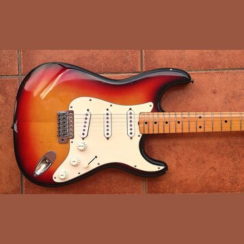 Elektrická kytara s tělem v červeno-žlutém sunburst provedení a bílým pickguardem. Nástroj má tři snímače typu single-coil, jeden přepínač a tři bílé…