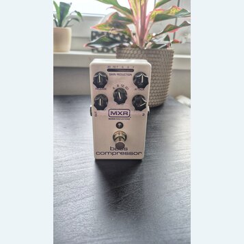 Bílý kytarový pedál s nápisy MXR, BASS INNOVATIONS a bass compressor. Na horní straně je LED indikátor GAIN REDUCTION s hodnotami -20, -10, -7, -5, -…
